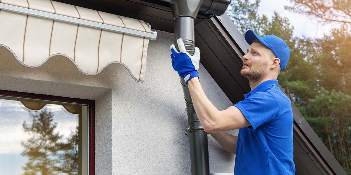 A man wearing a blue shirt and gloves is engaged in fixing a pipe, emphasizing the significance of gutter repair services.