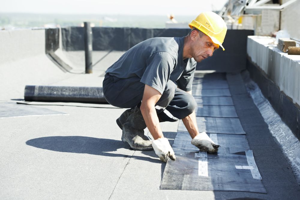 A construction worker in a yellow hard hat kneels on a rooftop, laying down black roofing material under bright sunlight.