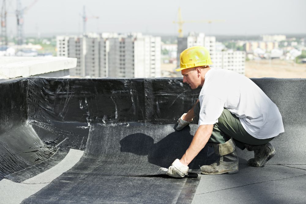 A construction worker in a yellow helmet applies roofing material on a building rooftop, with city buildings and cranes in the background.