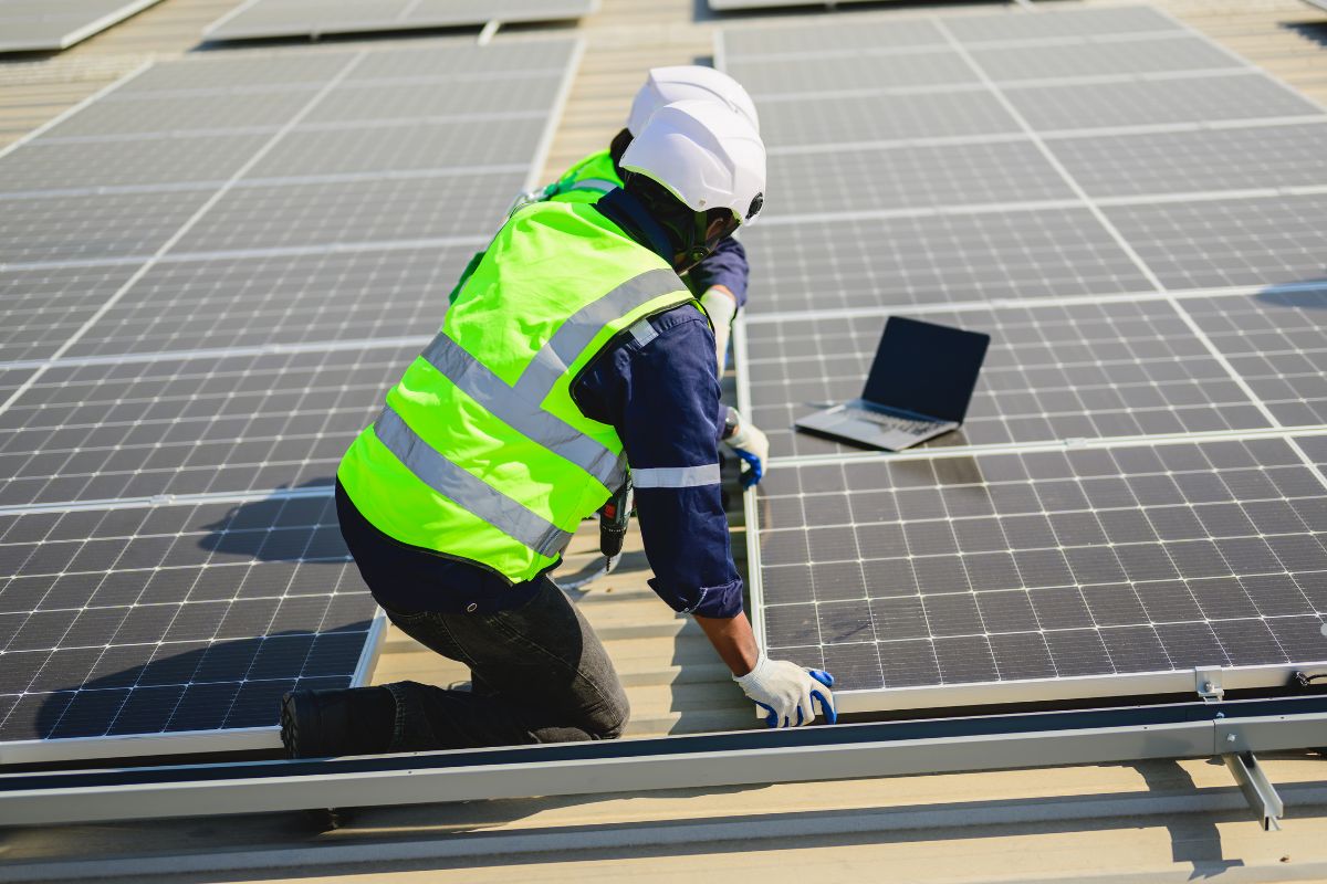 A worker in a safety vest and helmet installs solar panels on a rooftop, while a laptop is placed nearby for monitoring.