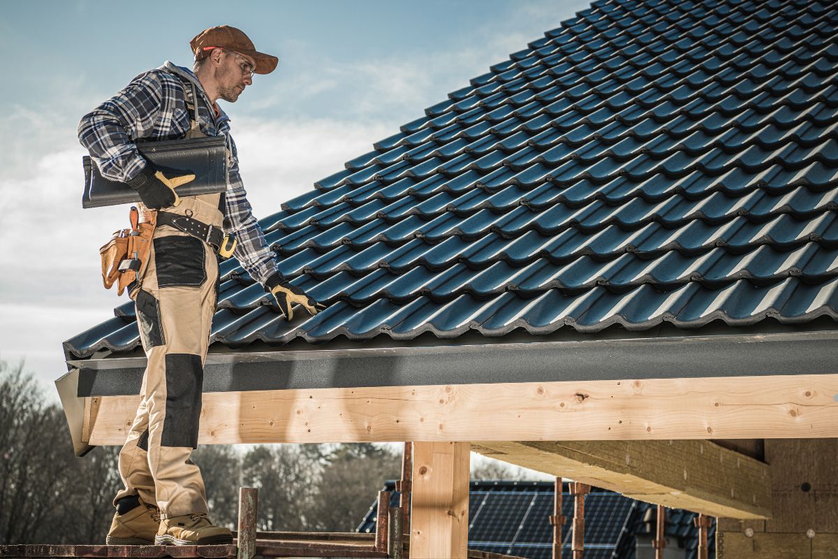 A worker in a plaid shirt and gloves stands on a sloped roof, adjusting tiles under a clear sky, showcasing construction activity.