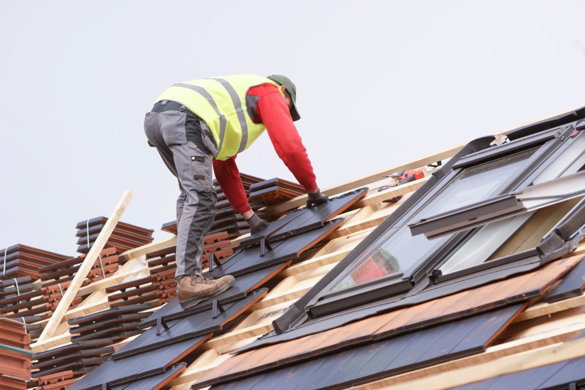 A worker installs solar panels on a roof, wearing a safety vest and gloves, amid roofing materials and a panoramic view.