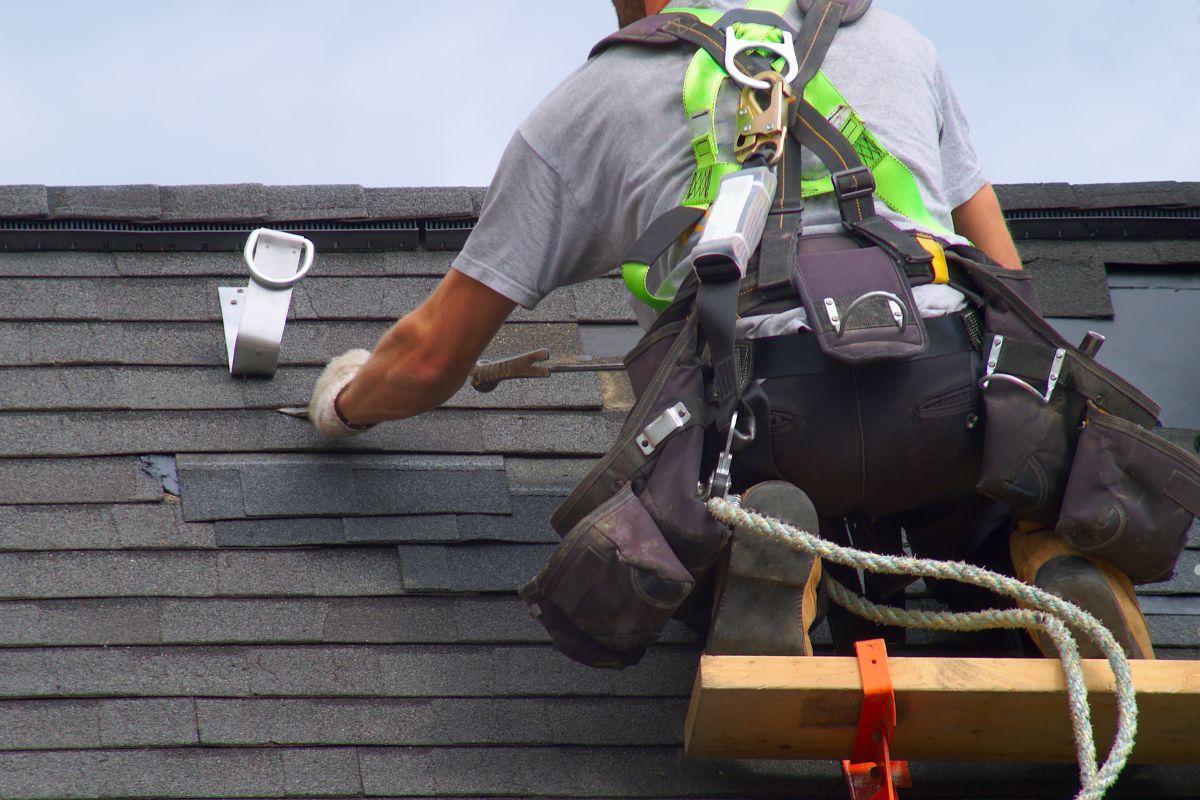 A roofer in safety gear working on shingles, using tools while secured with harnesses and ropes for safety.