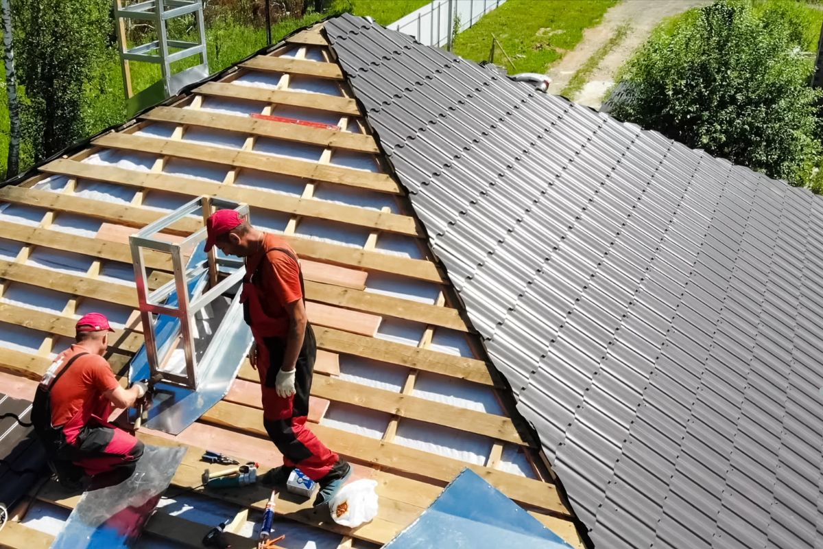 Two workers install roofing panels on a sloped metal roof, surrounded by wooden beams and green landscaping in bright sunshine.