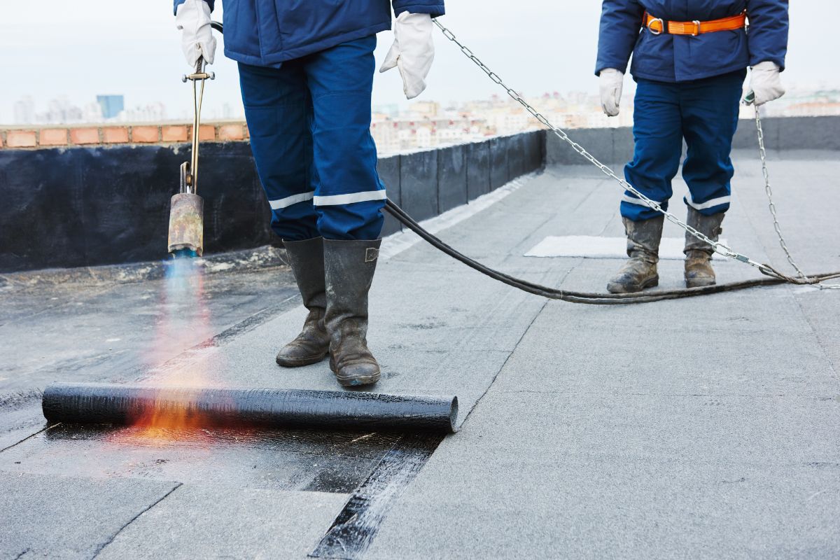 Workers in protective gear apply a heat torch to roofing material, sealing it with flame on a flat rooftop under a cloudy sky.