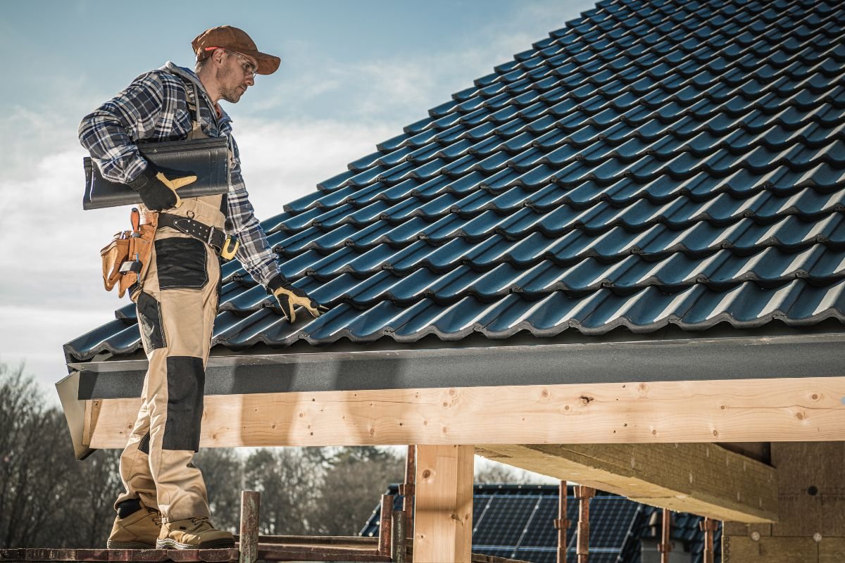 A construction worker in a plaid shirt and beige overalls stands on a roof, holding a binder and adjusting roof tiles under a blue sky.