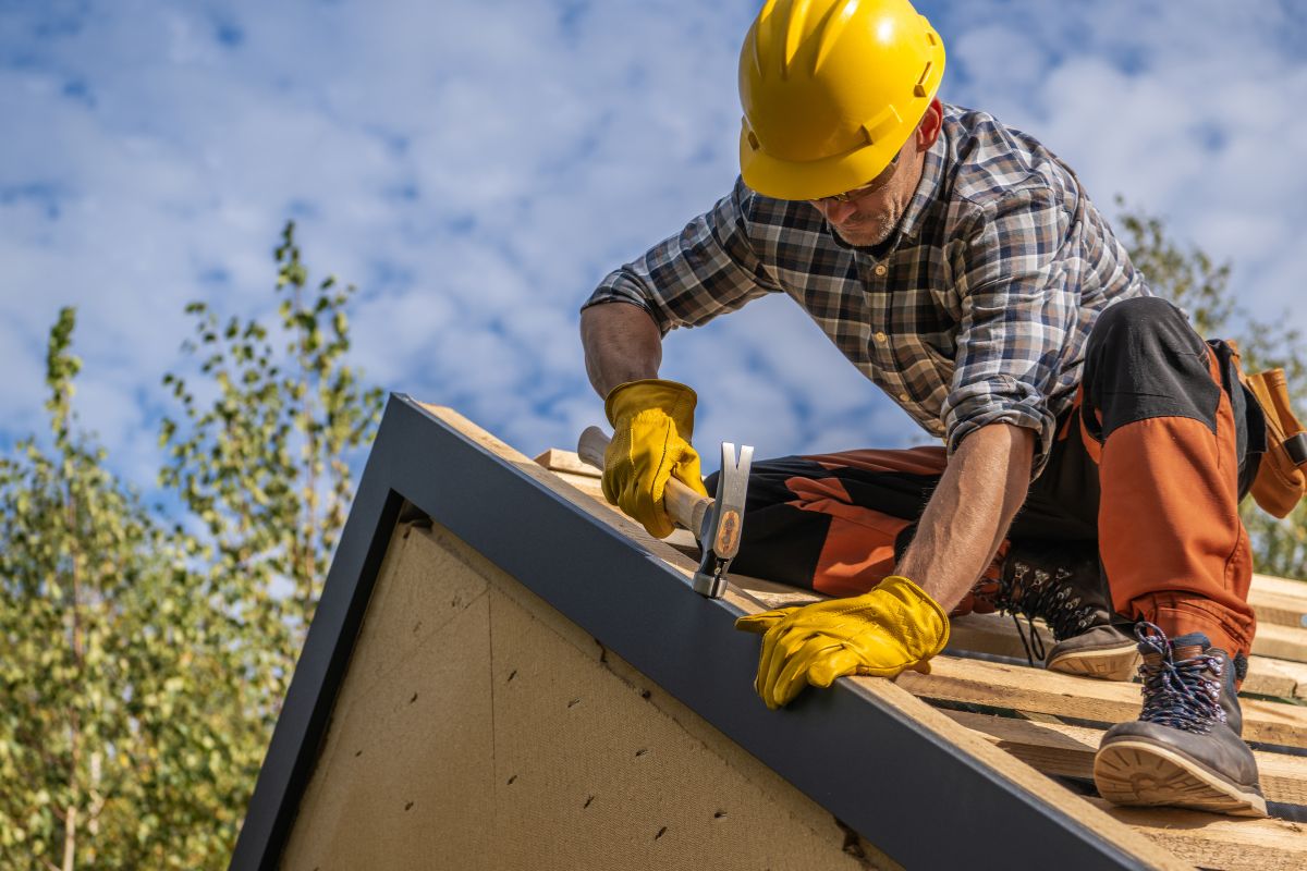 A construction worker wearing a yellow hard hat and gloves, using a hammer on a roof under a bright blue sky with scattered clouds.