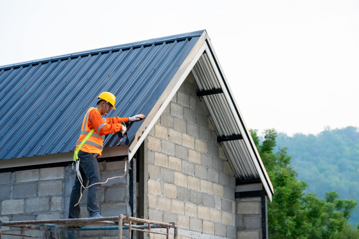 A construction worker in an orange safety vest and yellow hard hat installs roofing on a building under construction.